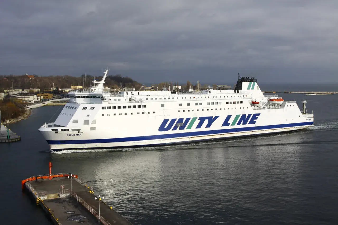 Biały prom Unity Line "Polonia" płynie po wodzie, w tle widać port i zachmurzone niebo.