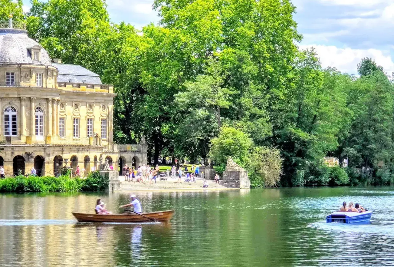 Ein sonniger Tag am Monrepos See. Menschen rudern in einem Boot, während im Hintergrund ein prächtiges Gebäude und üppige Bäume zu sehen sind.
