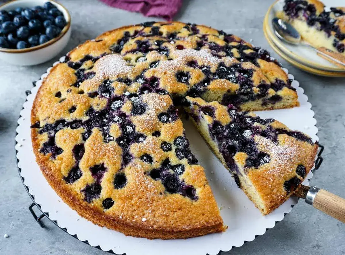 Ein saftiger Blaubeerkuchen, bestäubt mit Puderzucker, wird auf einem Teller serviert. Daneben eine Schüssel voller frischer Blaubeeren.