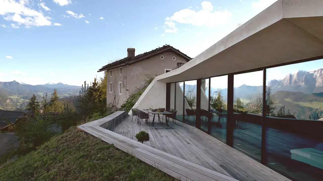 Modernes Haus in Hanglage mit Panoramafenstern und Terrasse, daneben ein altes Bauernhaus, umgeben von Berglandschaft.