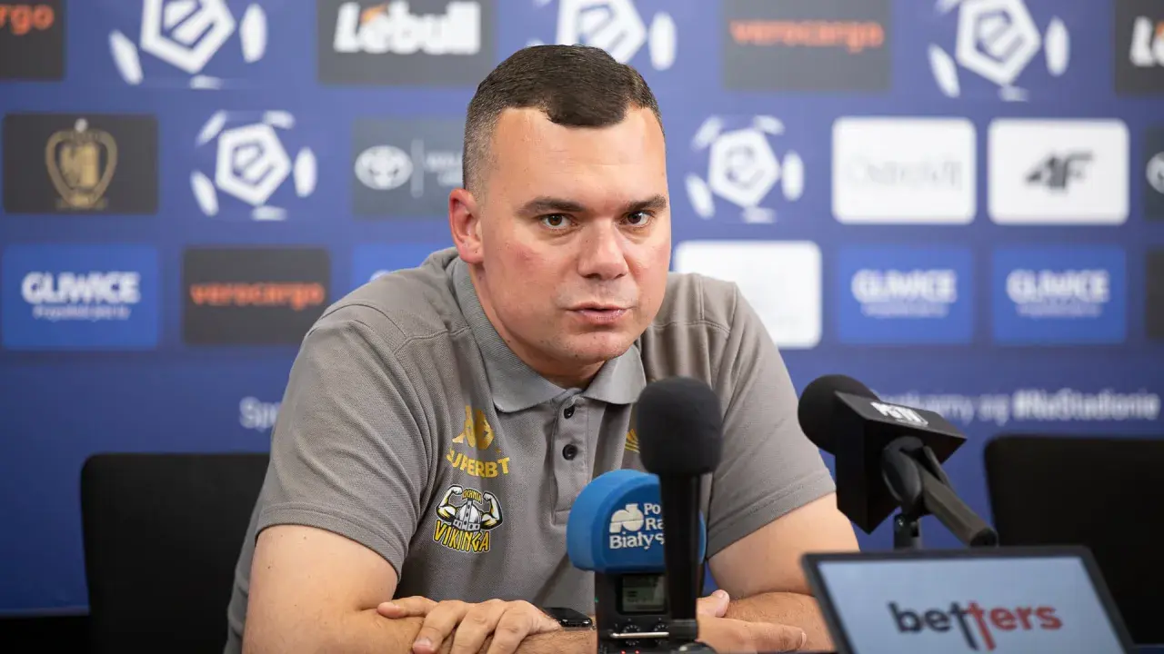 Trener Jagiellonii w szarej koszulce z logo klubu, siedzi przy mikrofonach podczas konferencji prasowej.