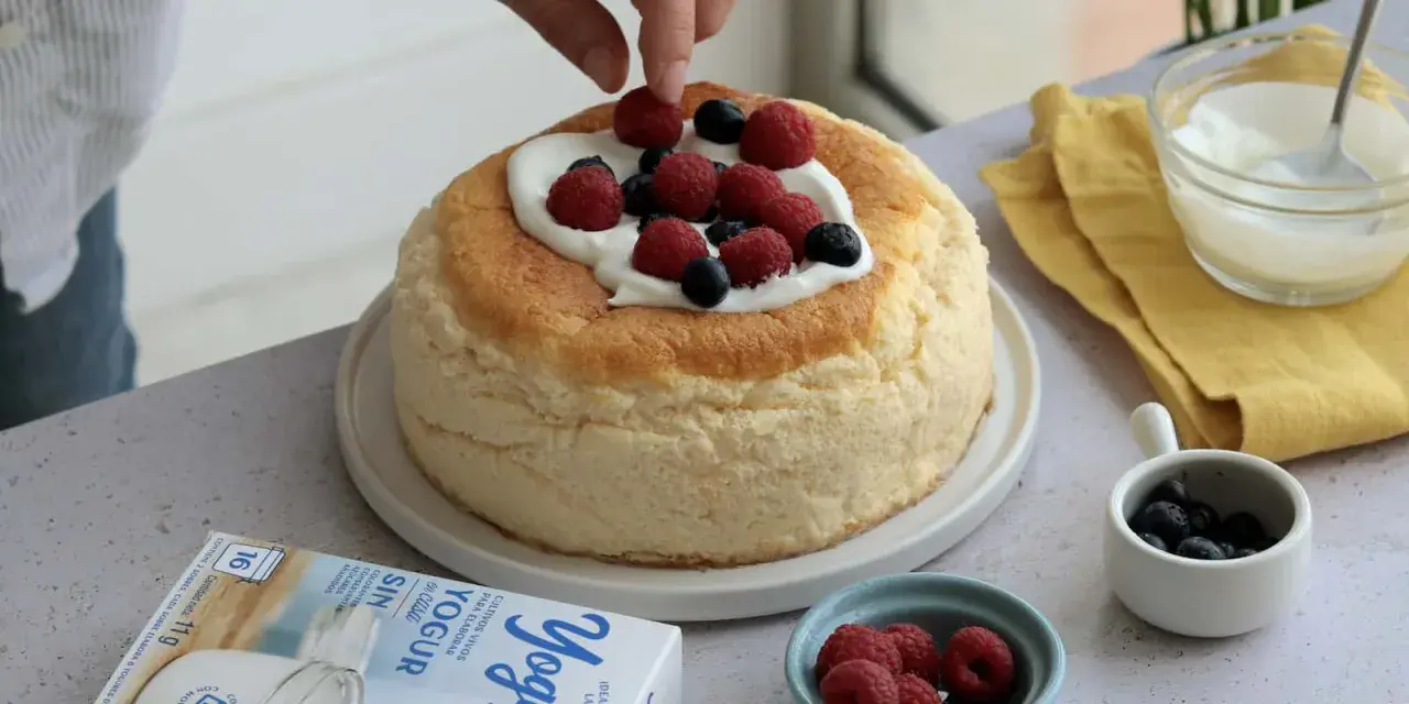Tarta de queso esponjosa decorada con frutos rojos y nata. Se ve una caja de yogur griego al lado.
