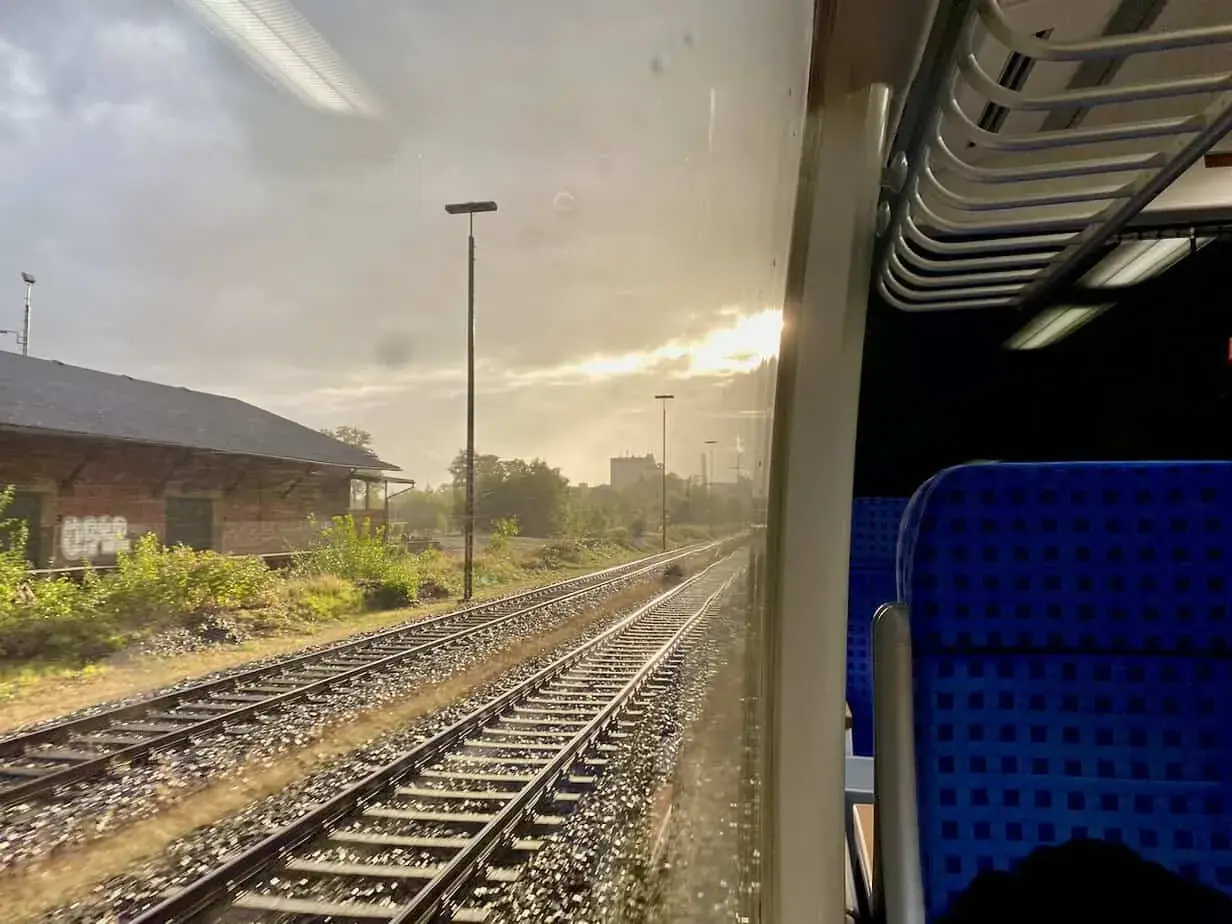 Blick aus dem Zugfenster auf Gleise und ein altes Backsteingebäude. Die Sonne bricht durch die Wolken. Man fragt sich, wie viel kostet eine Zugfahrt.