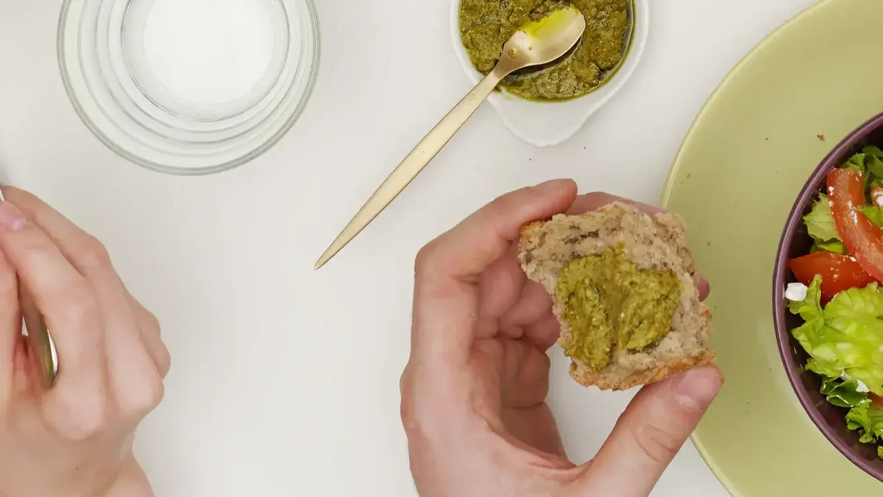 Main d'une personne tenant un morceau de pain avec du pesto, à côté d'une salade.