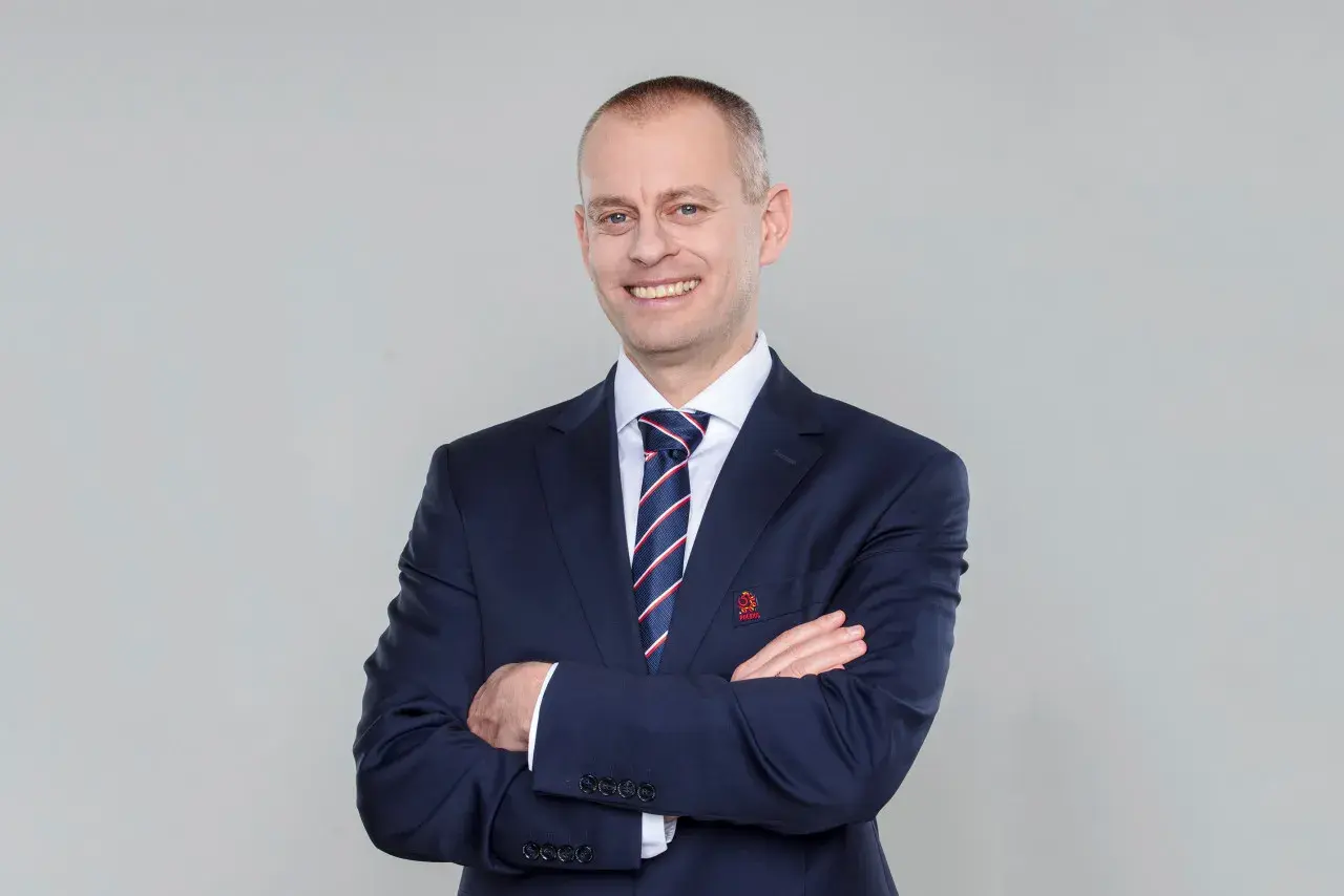 Marcin Włodarski, trener, w granatowym garniturze i krawacie, z rękami skrzyżowanymi na piersi, uśmiecha się do obiektywu.