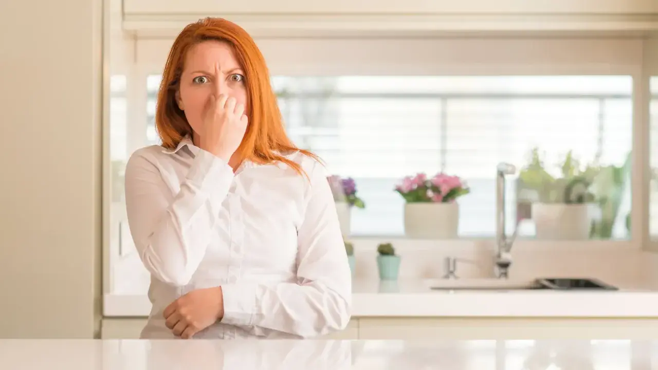 Mujer pelirroja se tapa la nariz, con cara de asco, por unos gases con olor a huevos podridos en la cocina.