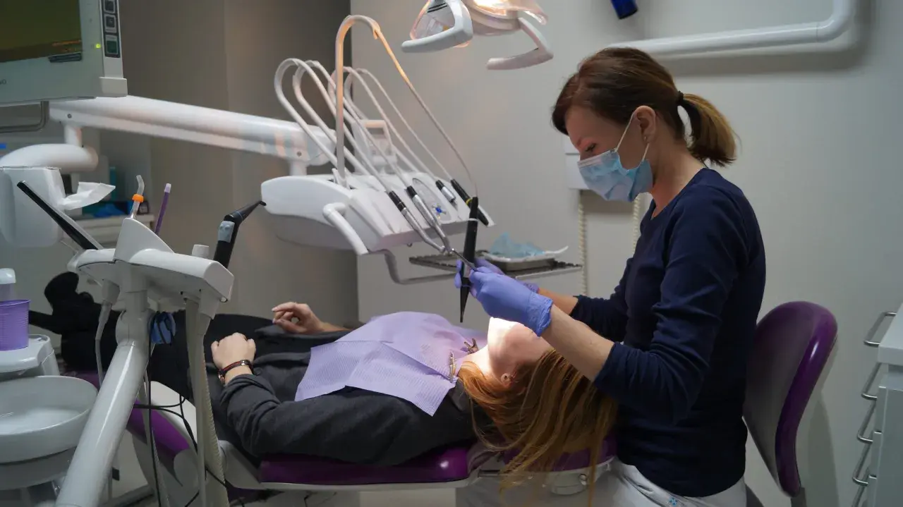 Dentystka w masce i rękawiczkach pracuje przy pacjencie. W gabinecie widoczny sprzęt do stomatologii.