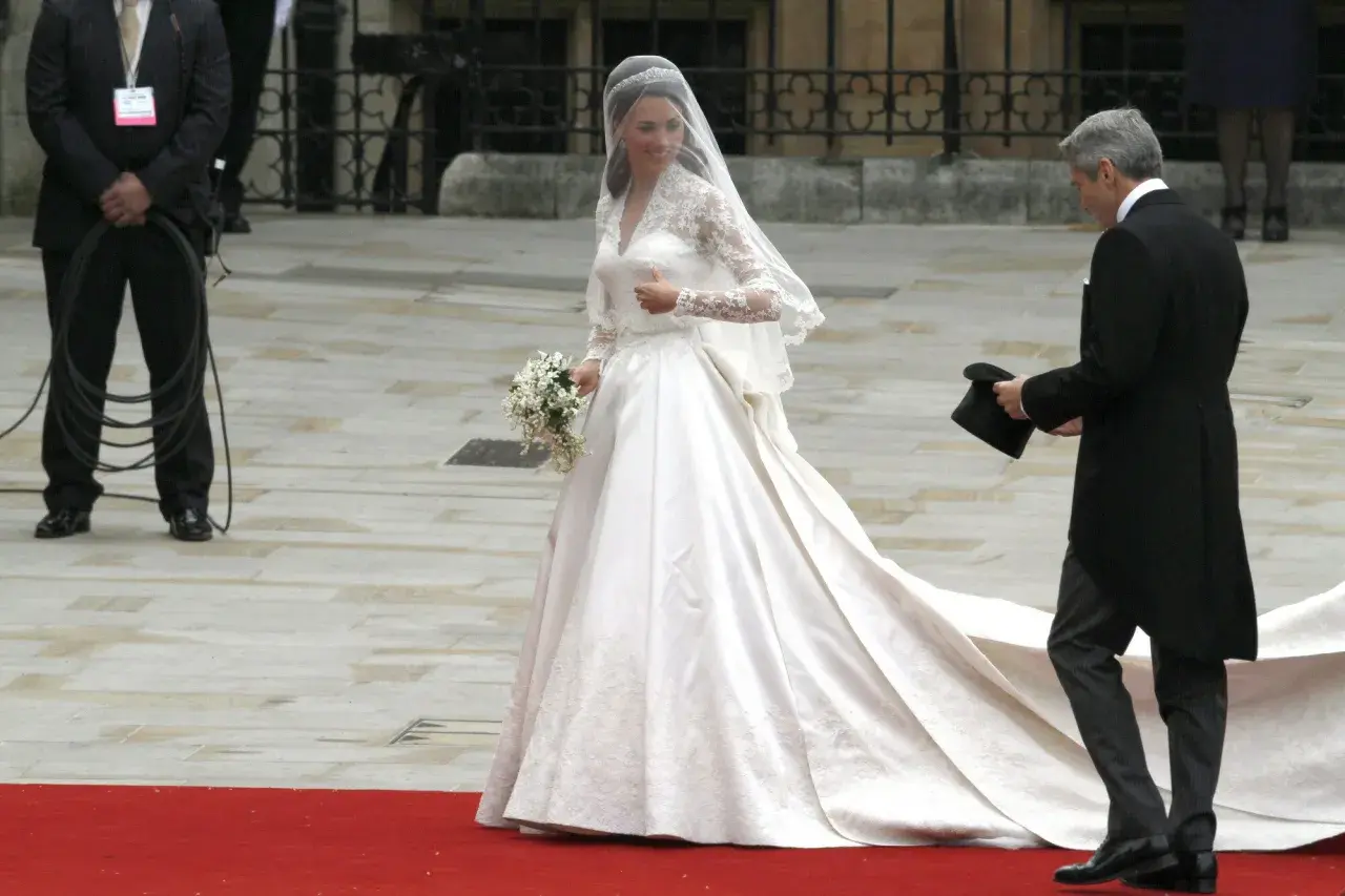 Kate Middleton w drugiej sukni ślubnej, z welonem i bukietem, idzie po czerwonym dywanie. Obok niej mężczyzna w żakiecie trzyma cylinder.