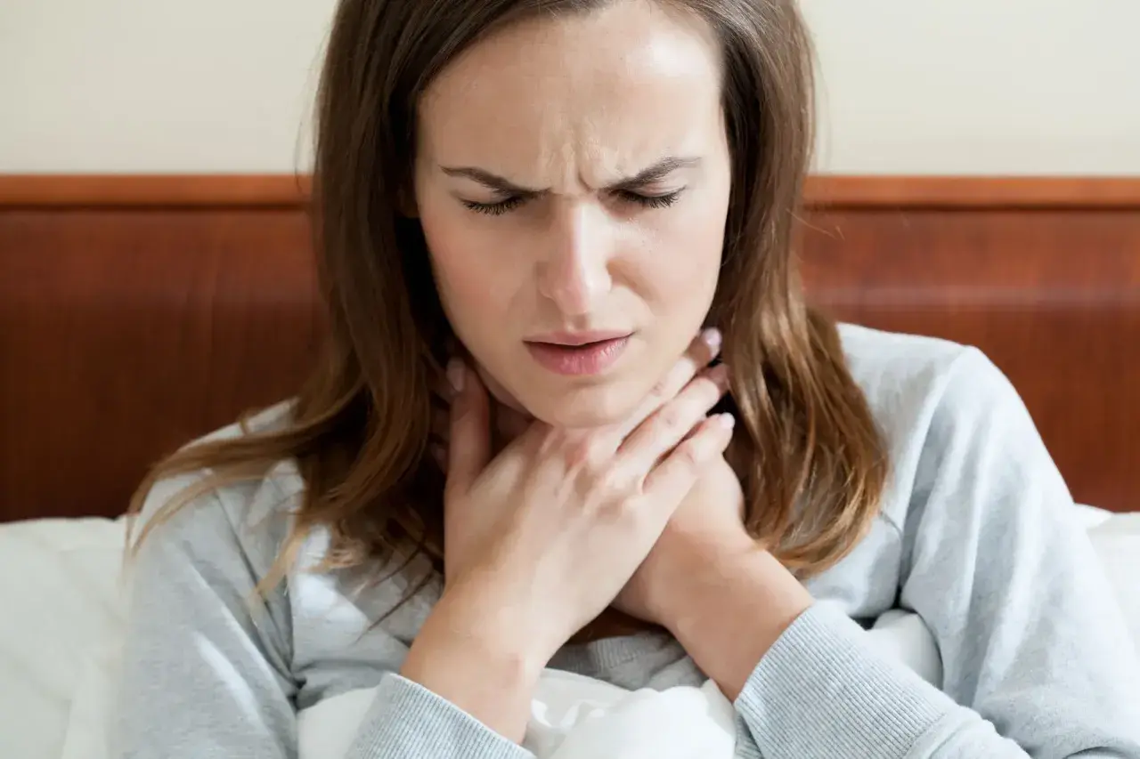 Donna con mani sul collo, espressione sofferente, forse per difficoltà a deglutire o ansia.