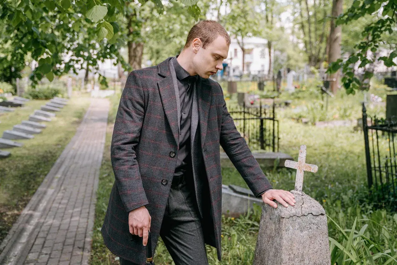 Mężczyzna w żałobnym stroju pochyla się nad kamiennym nagrobkiem z krzyżem na cmentarzu.