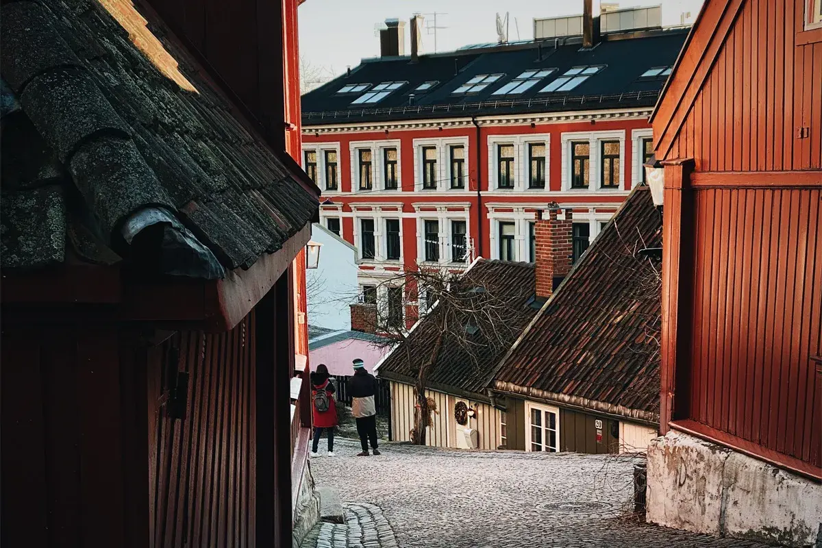 Två personer promenerar på en kullerstensgata i Oslo centrum, omgivna av charmiga trähus och en ståtlig röd byggnad.