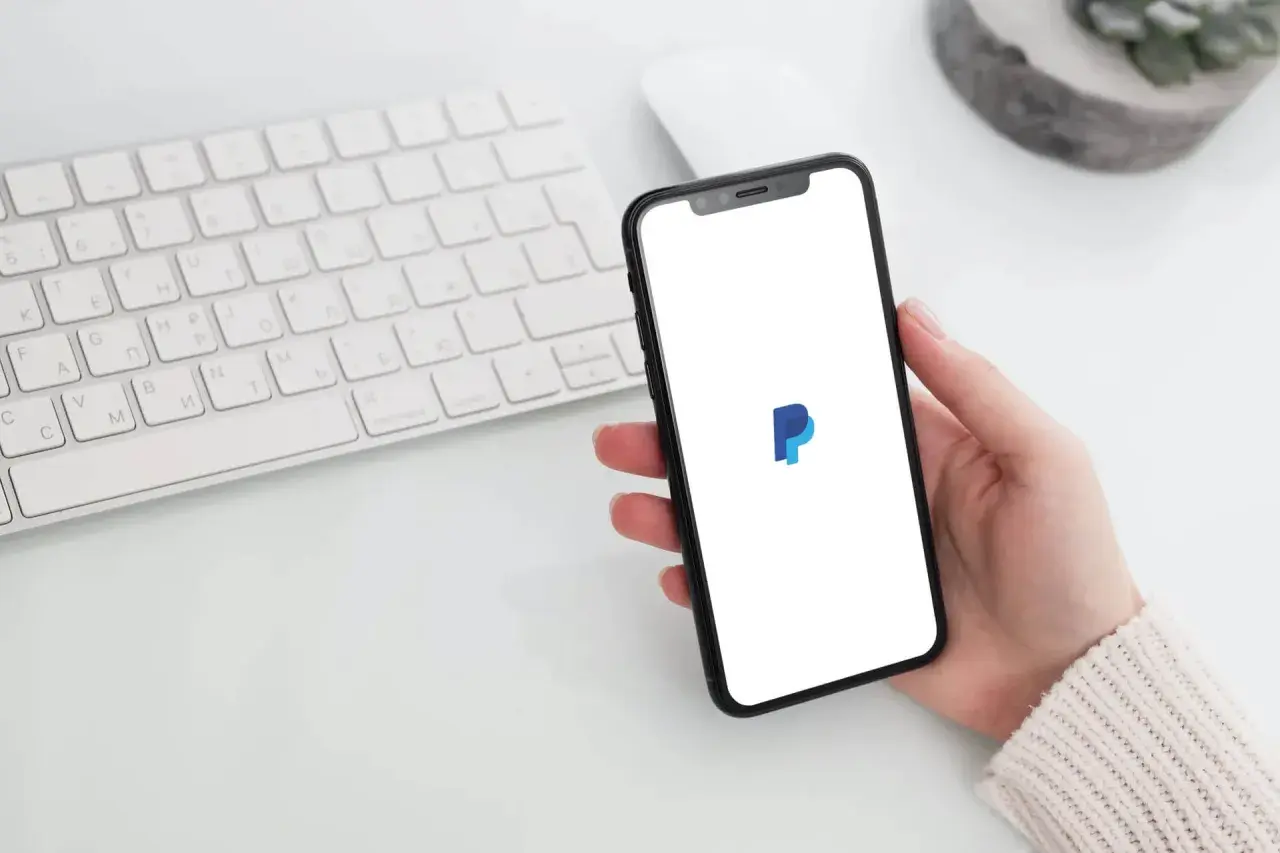Dłoń trzyma smartfon z logo PayPal. Czy PayPal jest darmowy? Sprawdź na telefonie, obok klawiatury.