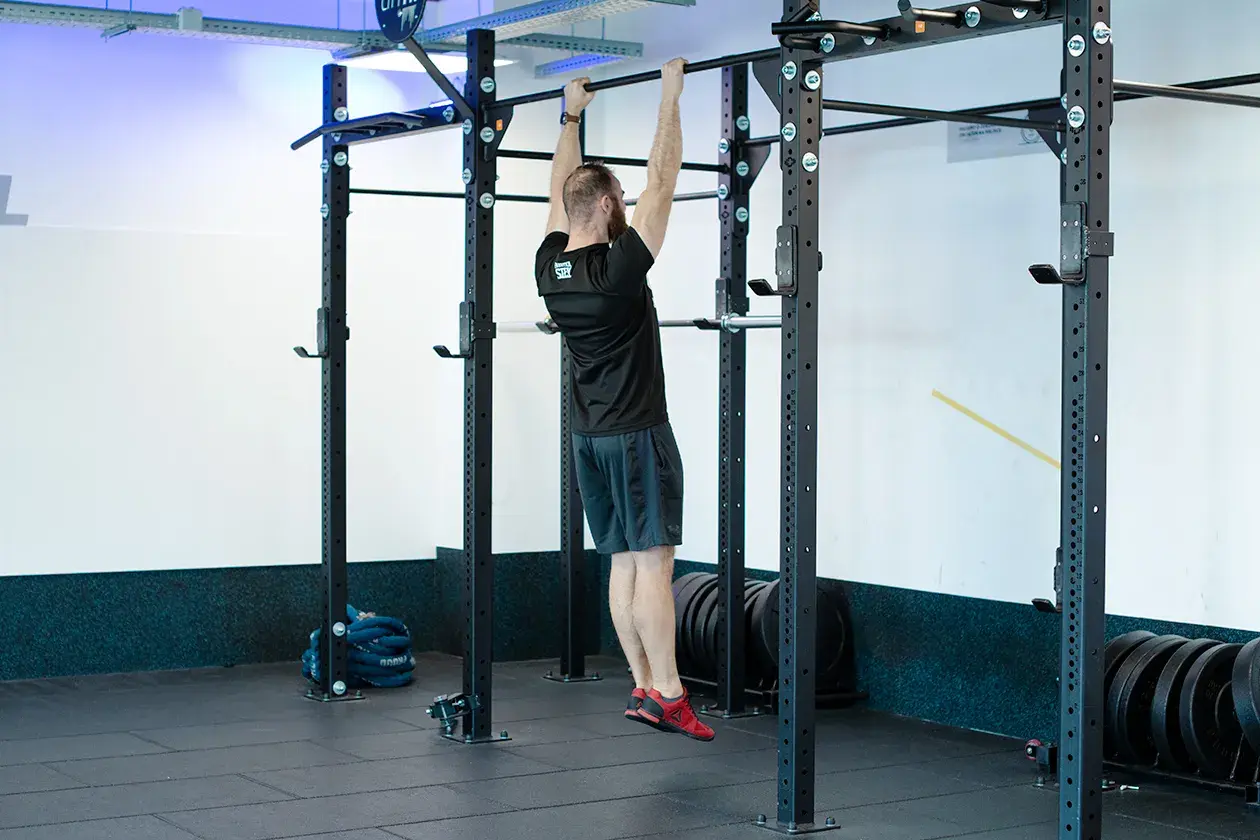Mężczyzna wykonuje scapular pull ups na drążku. Ćwiczy plecy i ramiona w siłowni.