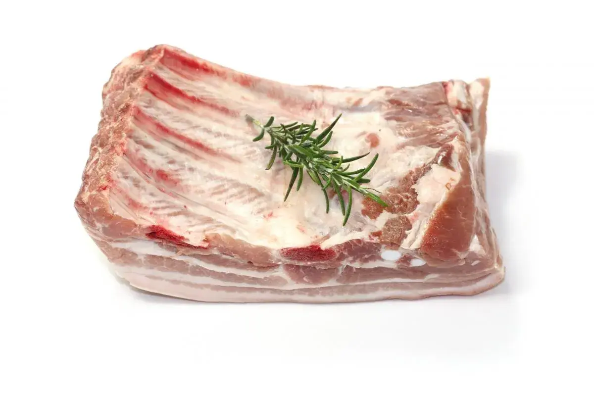 Un morceau de poitrine de porc frais, avec des côtes apparentes et une branche de romarin posée dessus, prêt à être cuisiné.