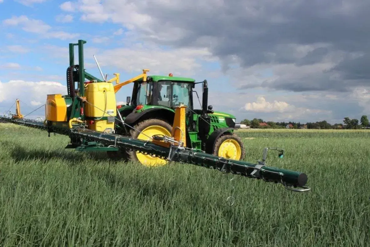 Zielony traktor z opryskiwaczem polowym, gotowy do pracy z adiuwantem, na tle pola i chmur.