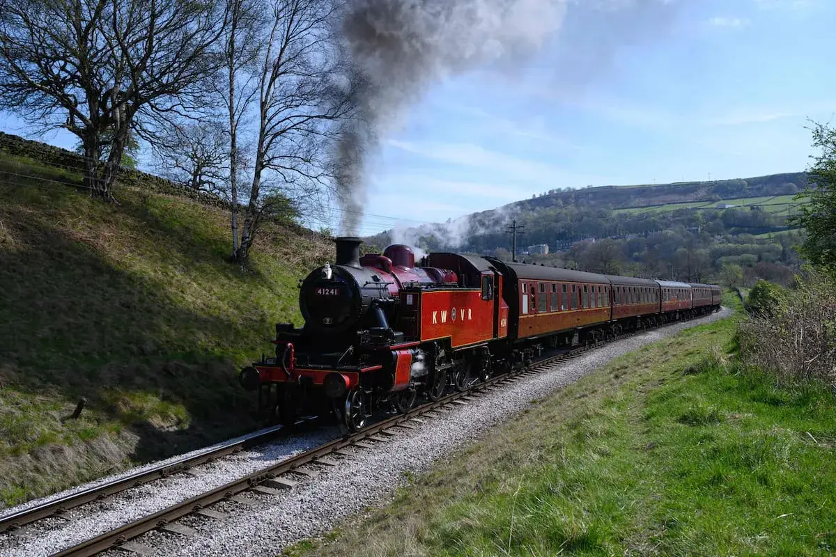 Un tren de vapor rojo y negro, con humo saliendo de su chimenea, recorre vías rodeadas de verde. Es como soñar con trenes antiguos en un paisaje idílico.