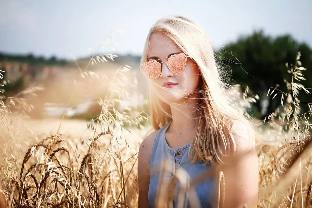 Dziewczyna z piaskowym blond włosami w okularach przeciwsłonecznych stoi w złotym polu pszenicy.