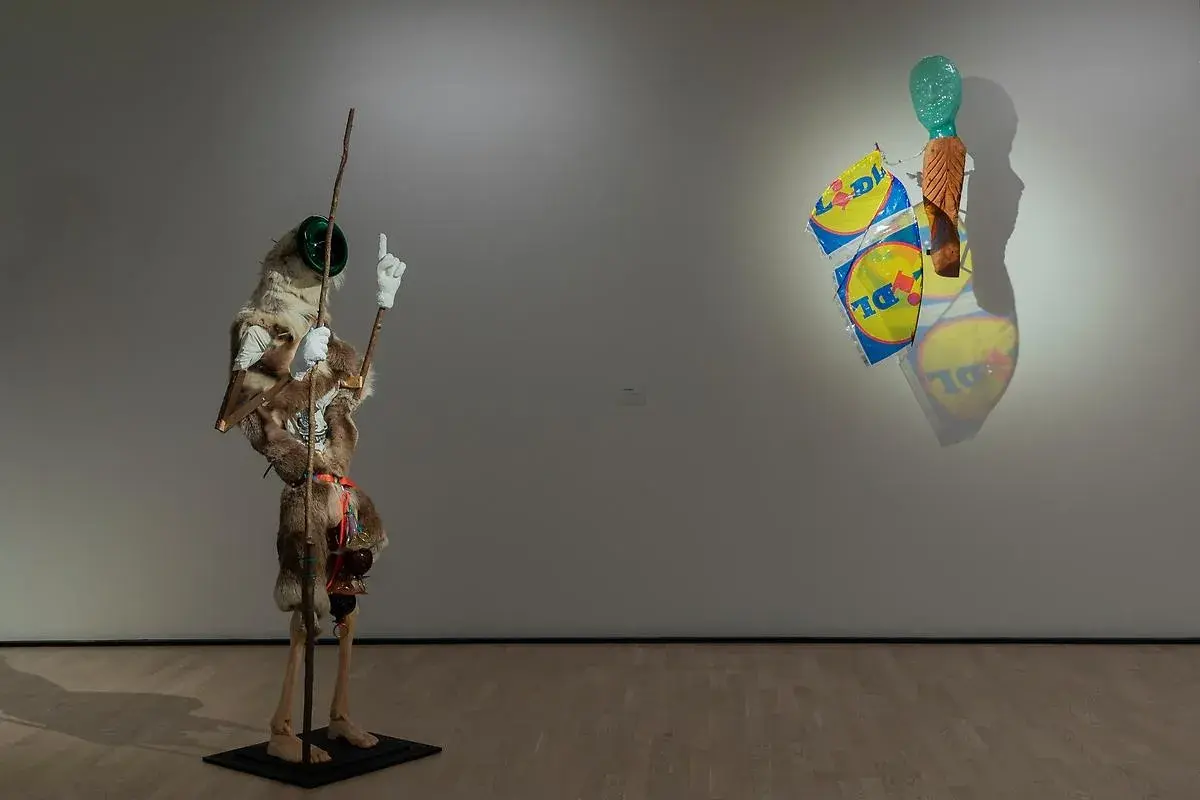Konsthallar Sverige: En surrealistisk figur med päls och en pinne står bredvid en väggskulptur med Lidl-påsar och ett grönt huvud.