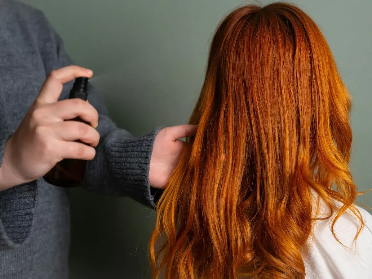 Persona rocía un producto en el cabello fino y quebradizo de color naranja rojizo para darle brillo y protección.