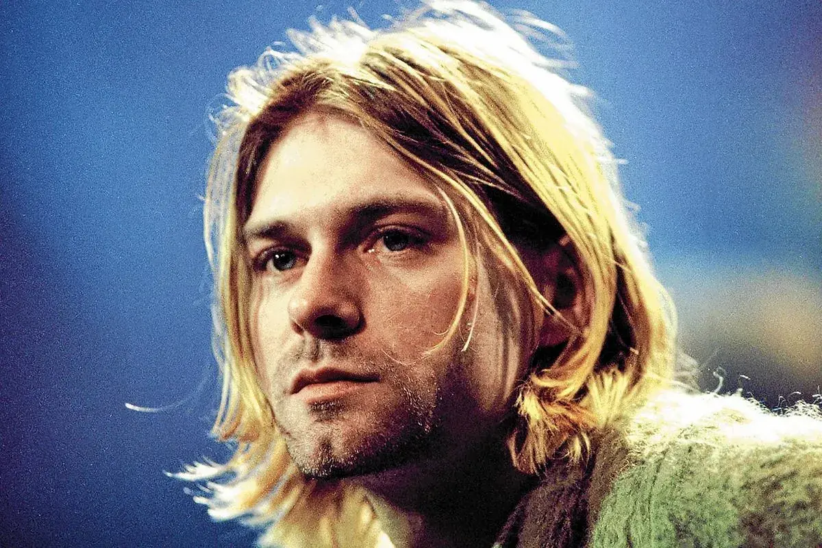 Kurt Cobain, ikona zespołu Nirvana, z charakterystycznymi blond włosami i niebieskimi oczami, na tle niebieskiego tła.