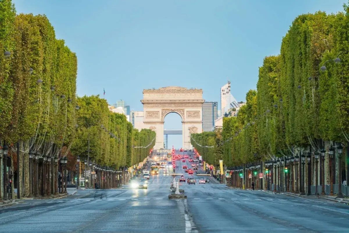 Aleja **Champs-Élysées** w Paryżu z Łukiem Triumfalnym w oddali. Drzewa tworzą zielony tunel, a ruch samochodowy płynie w stronę monumentu.