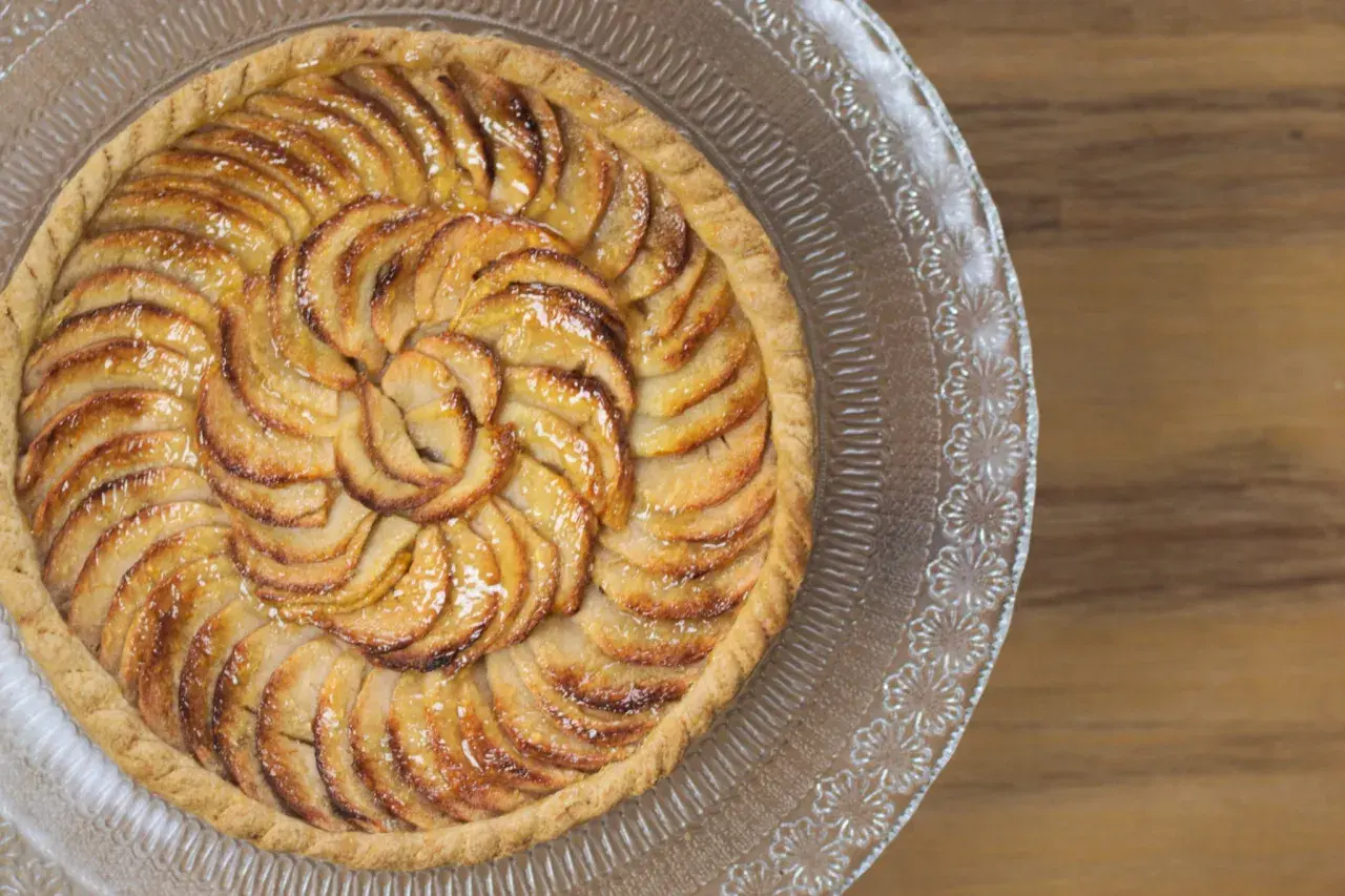 Un magnifique appareil tarte aux pommes, aux pommes caramélisées disposées en spirale sur une pâte dorée, sur un plat en verre décoré.