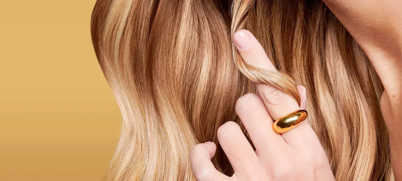 Capelli biondi con riflessi dorati, una mano con anello sfiora una ciocca. Il riflessante copre i capelli bianchi, donando luminosità.