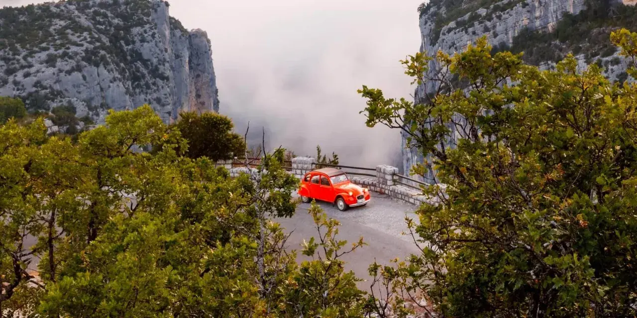 Une petite voiture rouge se gare sur la route des Gorges du Verdon, offrant une vue spectaculaire sur les falaises et la brume.