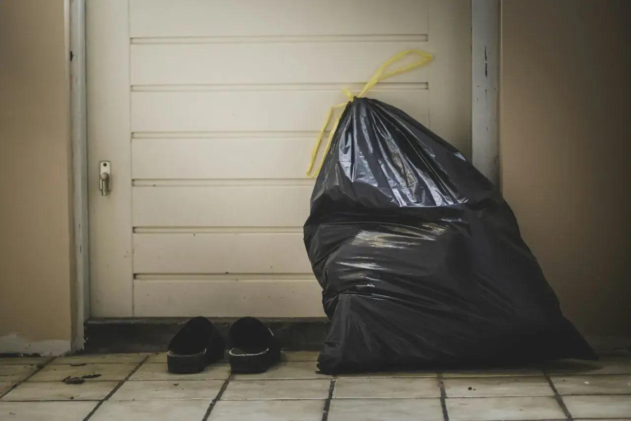 A full black trash bag and a pair of slippers sit by a closed door, symbolizing the need for lean budgeting to discard unnecessary expenses.