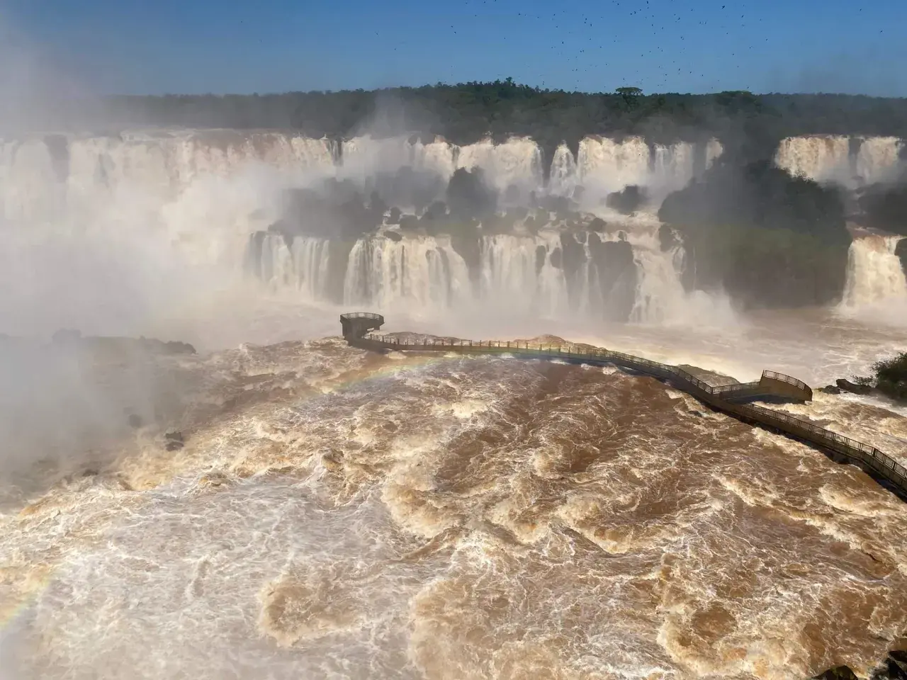Mächtige Iguazú-Wasserfälle stürzen in schlammbraunes Wasser. Ein Steg führt durch den Sprühnebel, ein Regenbogen funkelt.