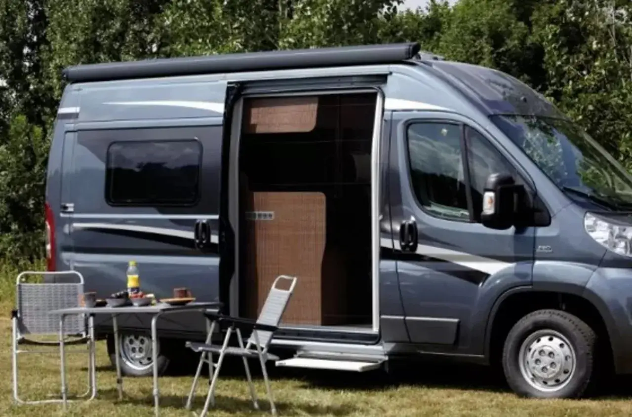 Un camping-car gris avec une moustiquaire plissée de porte ouverte. Une table et des chaises sont installées dehors.