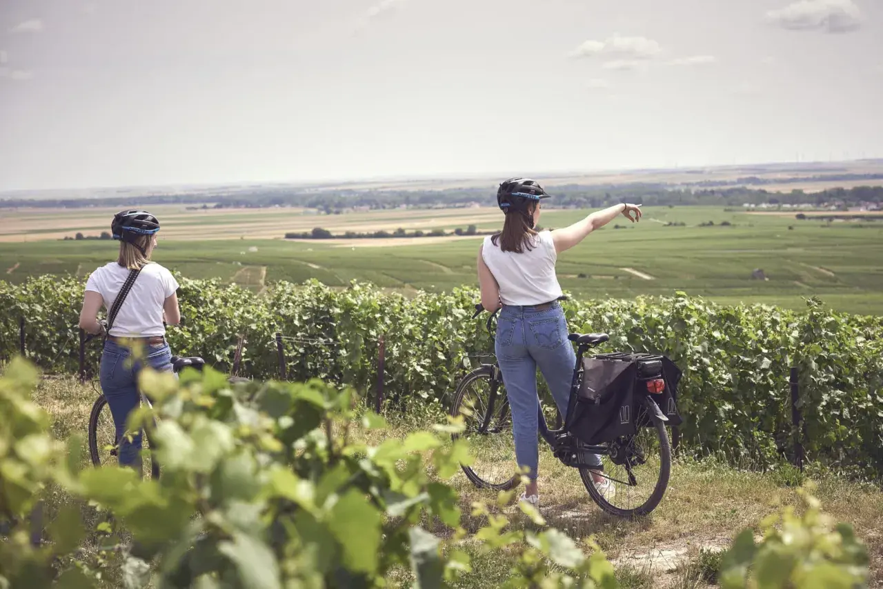 Two women on a champagne bike tour point towards rolling vineyards under a hazy sky.
