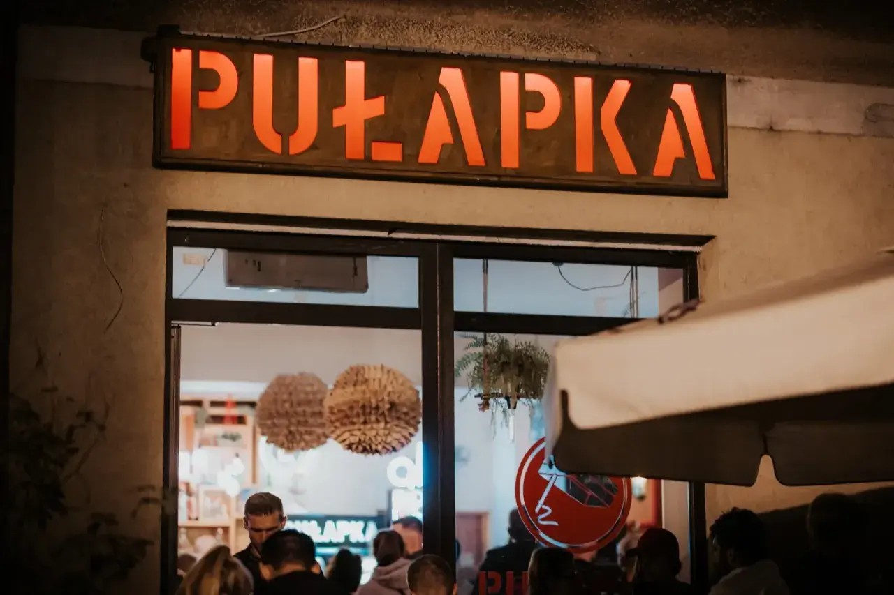 Neonowy napis "PUŁAPKA" nad wejściem do lokalu. W środku widać ludzi i dekoracje.