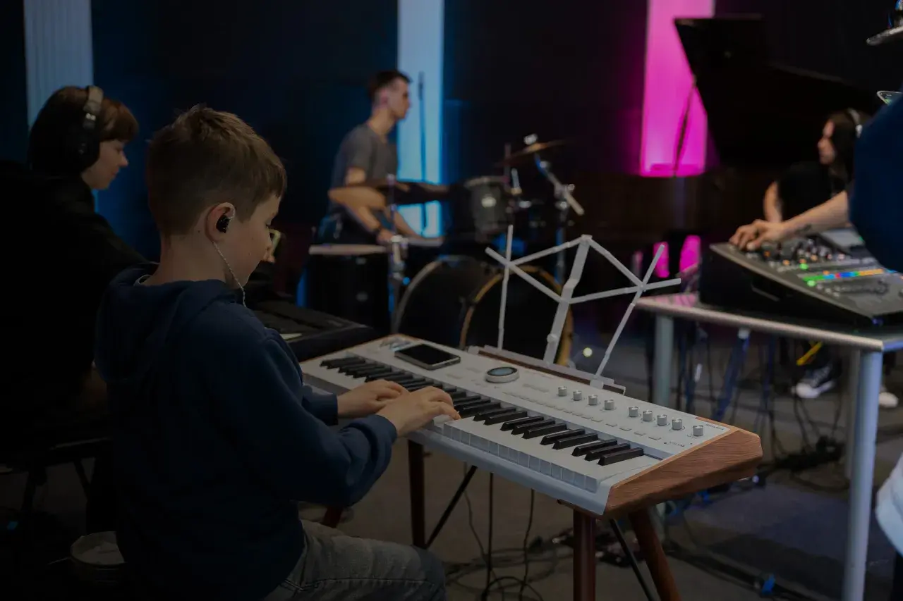 Chłopiec na lekcjach śpiewu ćwiczy grę na keyboardzie, podczas gdy w tle słychać perkusję i mikser dźwięku.