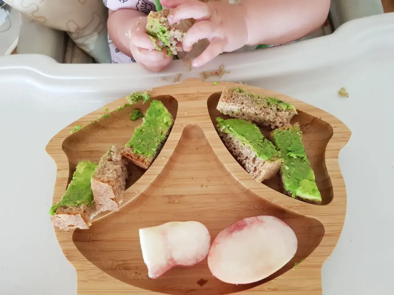 Babyhände greifen nach Avocado-Brotstücken auf einem Holzbrett. Ein 10 Monate altes Baby erkundet sein Essen.