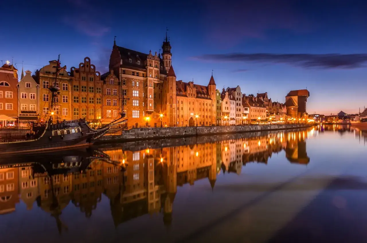 Gdańsk nocą, z zabytkowymi kamienicami i żaglowcem odbijającymi się w spokojnej wodzie Motławy.