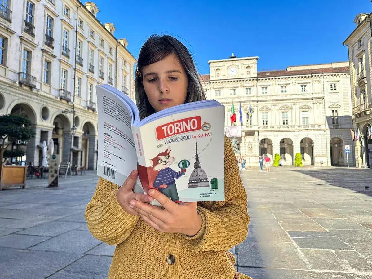 Bambina legge una guida "Torino con Viaggiapiccoli" in una piazza torinese. La libreria viaggi è perfetta per scoprire la città.