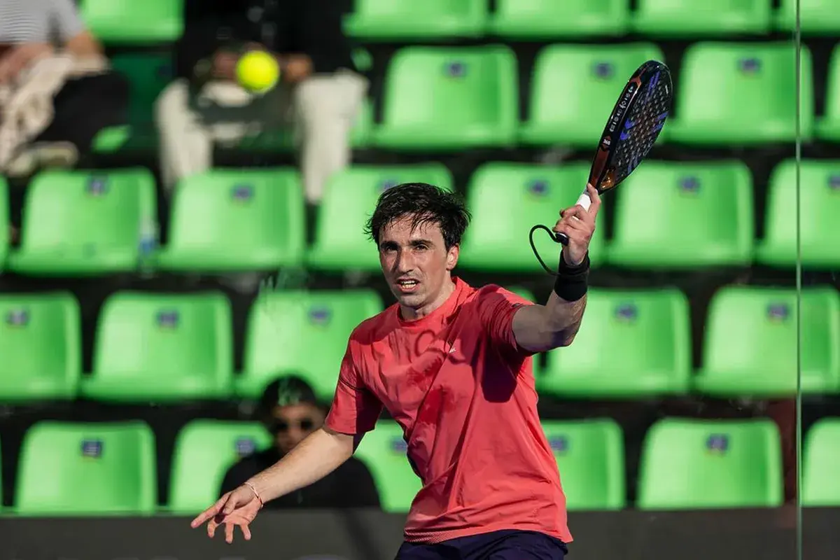Giocatore di padel in azione, con la racchetta sollevata e la palla in volo. Le tribune verdi sullo sfondo creano un contrasto vibrante.