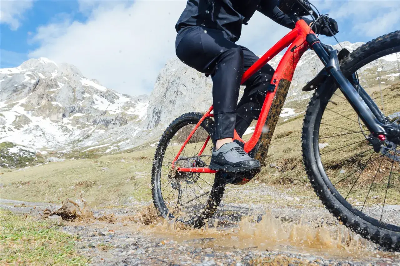 Kolarz na czerwonym rowerze trekkingowym pokonuje błotnistą kałużę na górskim szlaku.