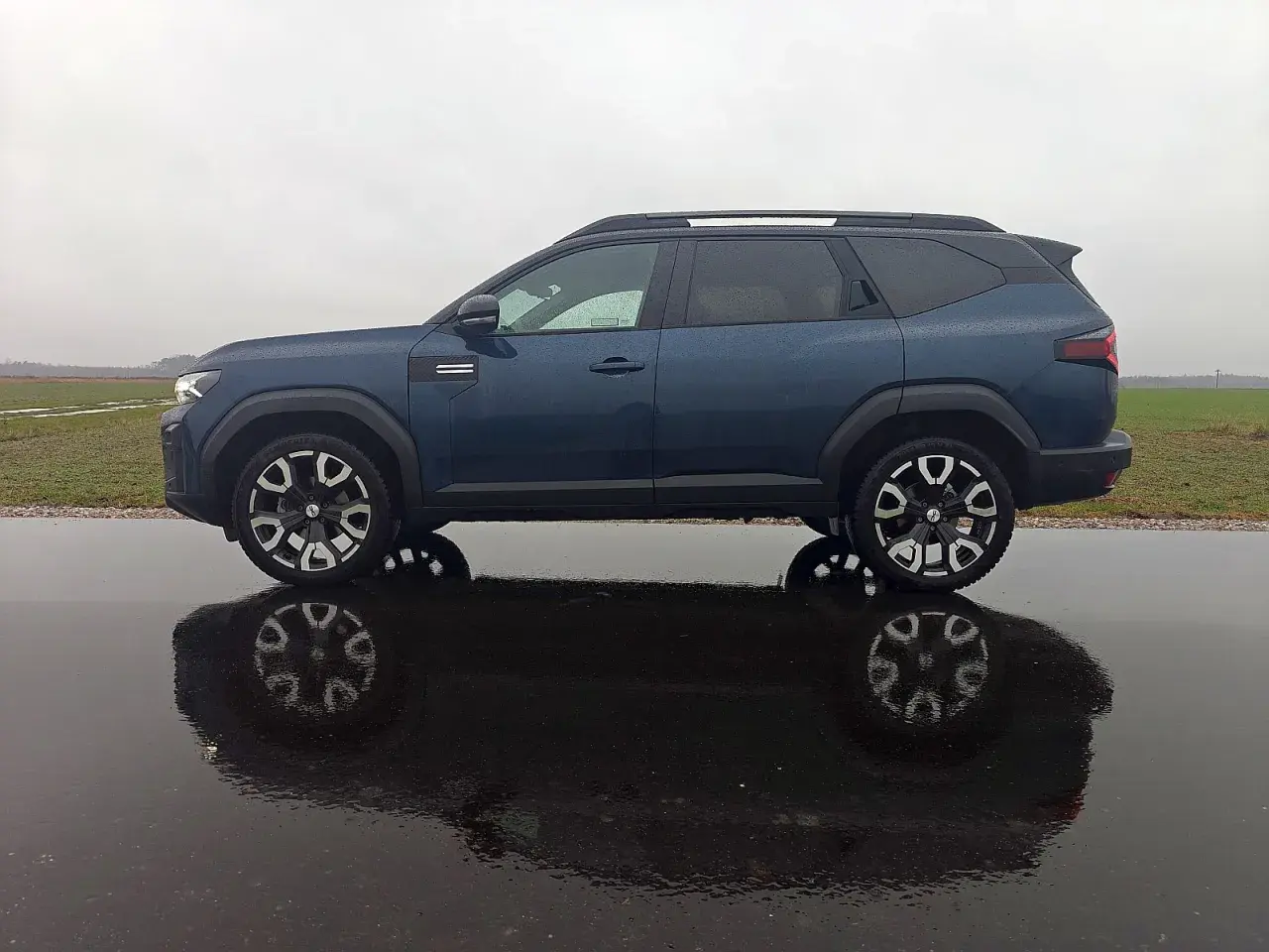 Dacia Bigster LPG w ciemnoniebieskim kolorze, zaparkowana na mokrej drodze, z odbiciem w kałuży.