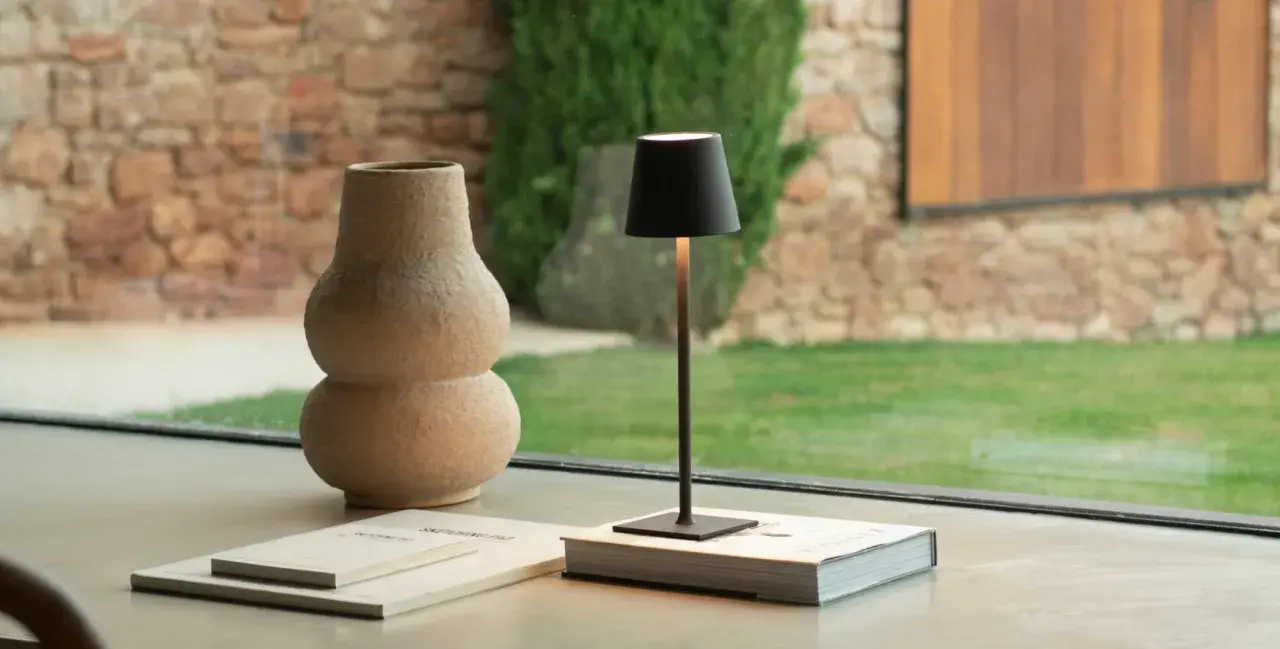Elegantes ideas de lámparas para tu hogar: una lámpara negra minimalista sobre un libro, junto a un jarrón texturizado y un jardín exterior.