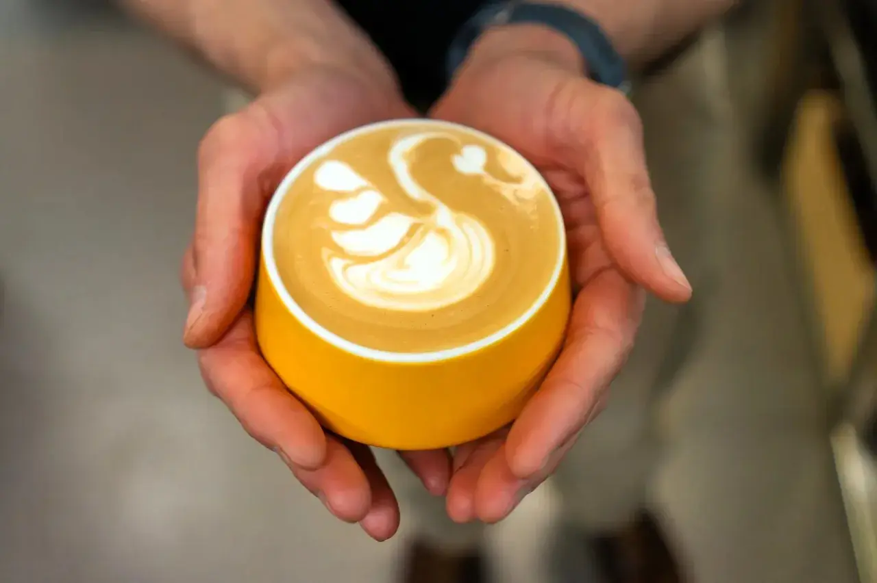 Dłonie trzymają żółtą filiżankę z kawą, na której artystycznie uformowano łabędzia. Zastanawiasz się, ile kofeiny ma espresso z ekspresu?