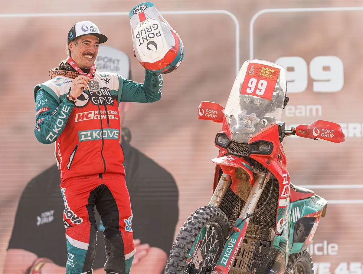 Piloto celebra victoria junto a su moto china Dakar. Casco y medalla en mano, luce orgulloso su equipación.