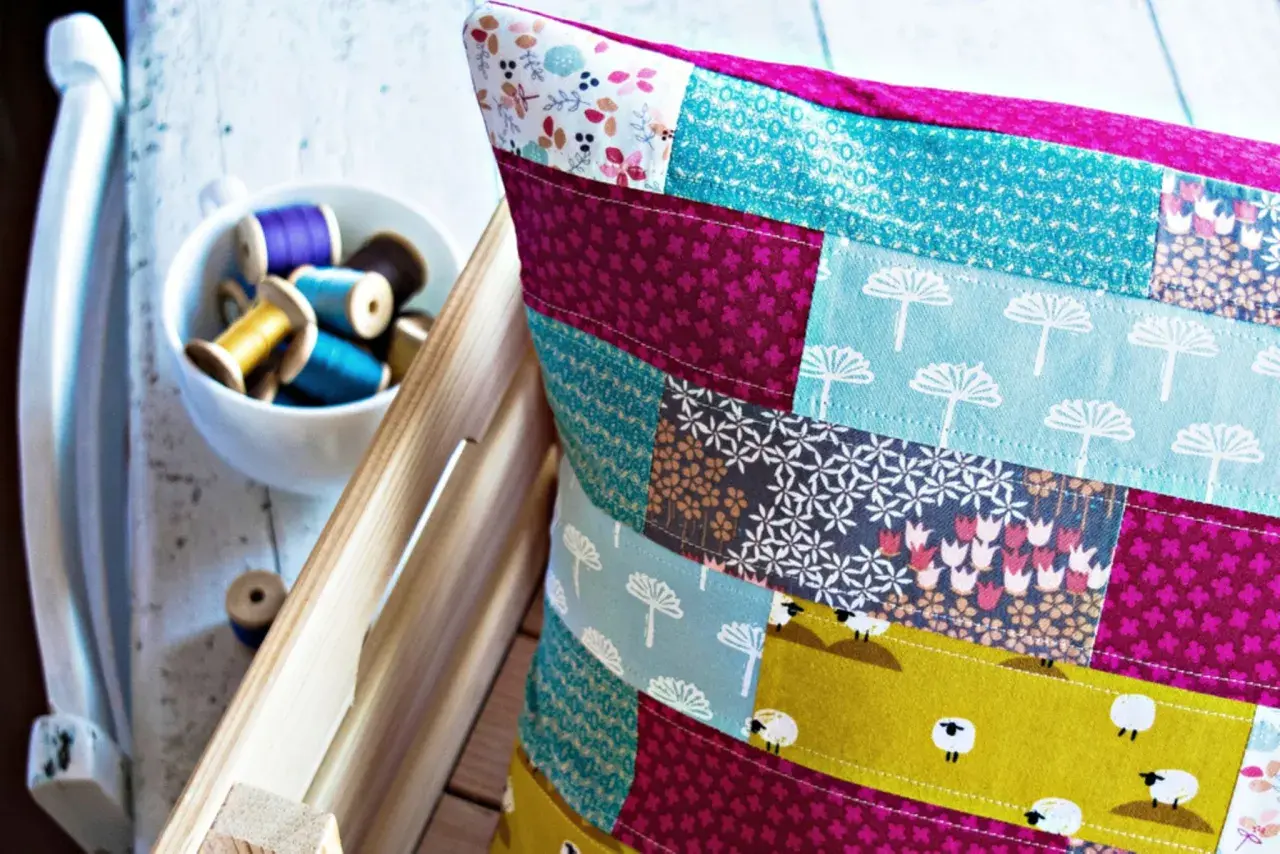 Buntes Patchwork-Kissen zeigt, wie man Stoffreste sinnvoll verwerten kann. Kleine Schafe und Blumenmuster zieren die Kissenhülle.