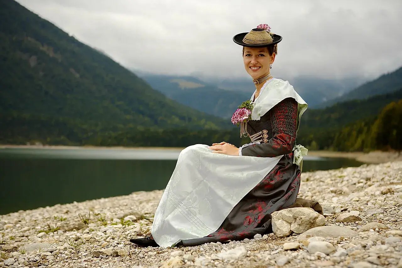 Frau in traditioneller Isarwinkler Tracht sitzt am Ufer eines Sees vor einer Bergkulisse.