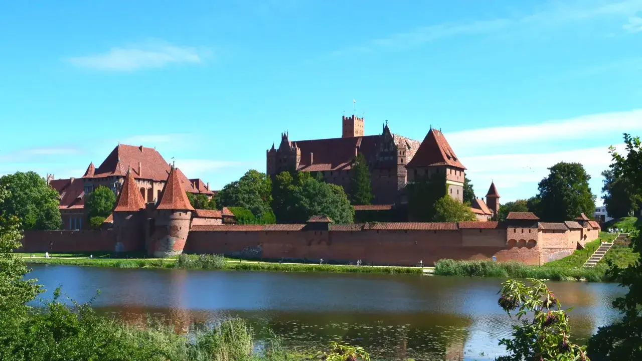 Potężny zamek Malbork z czerwonej cegły, otoczony murami obronnymi, góruje nad spokojną wodą.