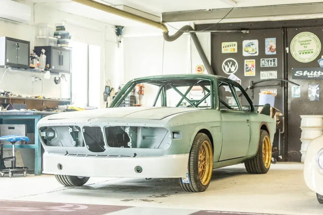 En grön BMW E9 i verkstad, redo för renovering. Guldiga fälgar och en imponerande bur syns tydligt.