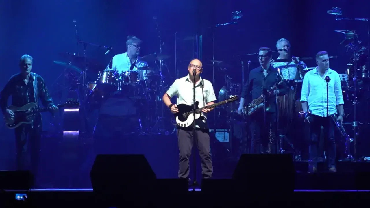 Mark Knopfler na scenie z zespołem podczas jednego z jego niezapomnianych koncertów. Muzycy grają na gitarach, saksofonach i perkusji.