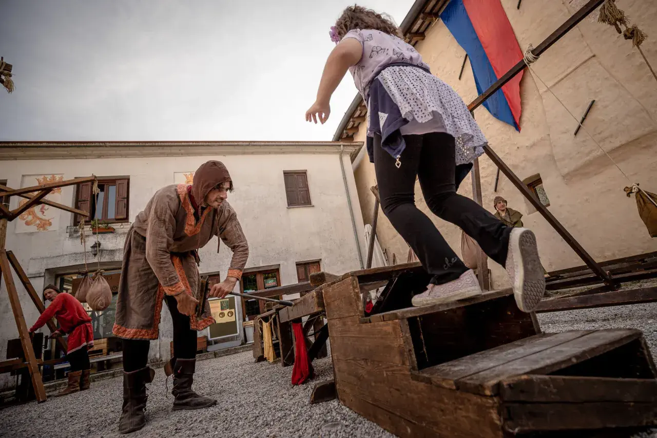 Bambini si divertono con giochi medievali. Un uomo in costume d'epoca aiuta una bambina a salire su una struttura di legno.