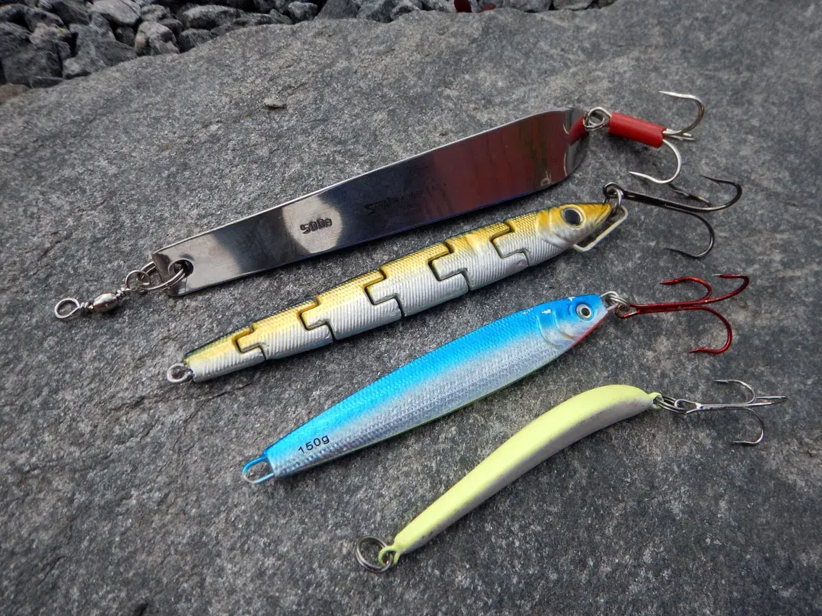 Vier Kunstköder für das Pilken vom Ufer liegen auf grauem Stein. Ein langer Silberpilker, ein gelb-silberner Wobbler, ein blauer Wobbler und ein gelber Gummifisch.